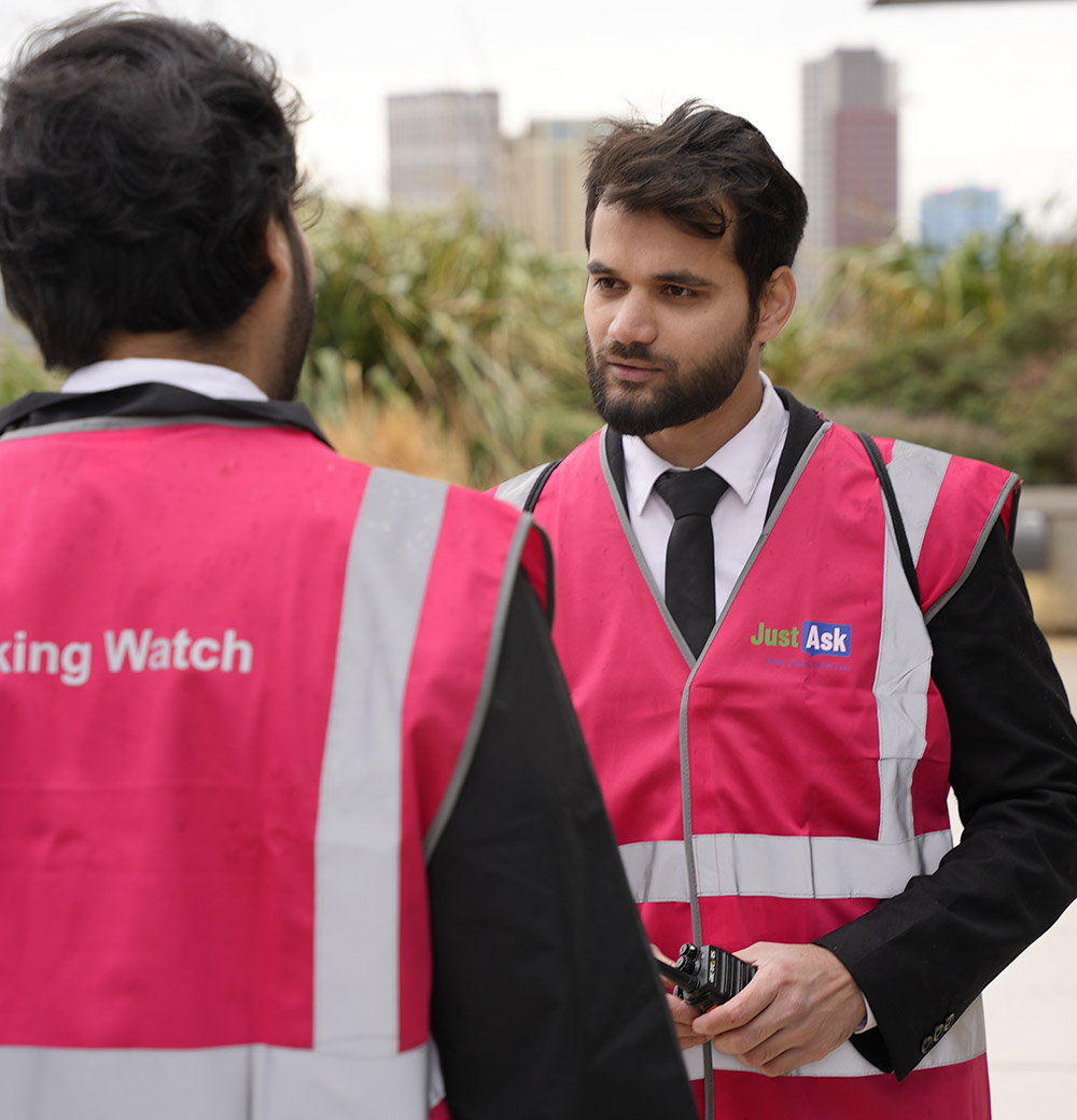 Two team members wearing Just Ask high-visibility vests discussing site operations during a waking watch inspection.