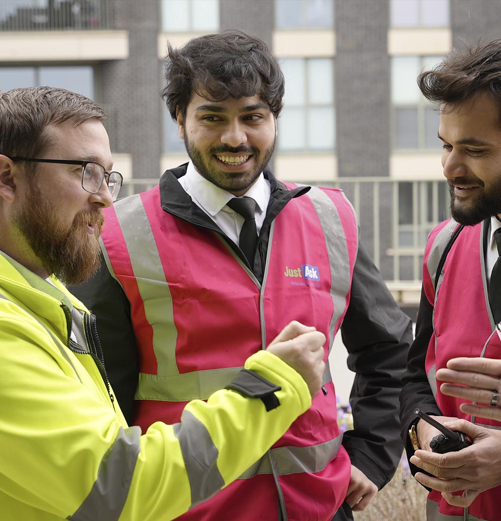 A JustAsk waking watch team in high-visibility vests discussing safety procedures outside a residential building, highlighting teamwork and collaboration during site patrols.